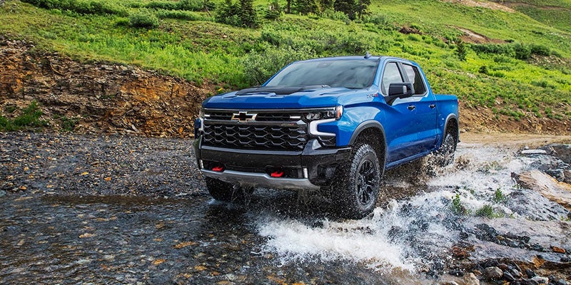Blue 2022 Chevrolet Silverado 1500 driving through a rocky stream with splashing water, set against a grassy hillside background.