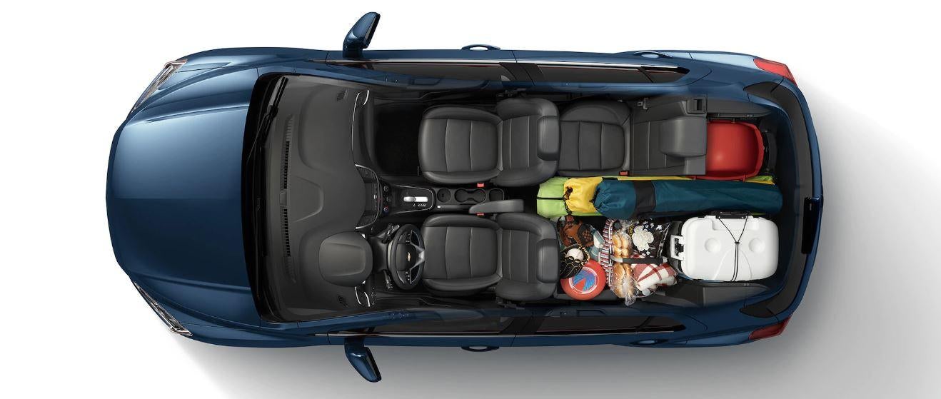 Top-down view of a blue 2025 Chevy Trax with the roof removed, showing black interior seats and cargo area packed with camping gear, including a tent, cooler, beach toys, and picnic supplies.