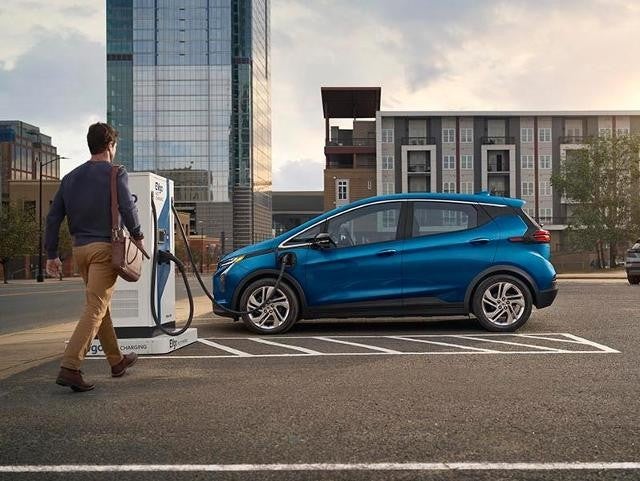 Man walking toward a blue electric Chevrolet Bolt EUV charging at a public charging station in a modern urban setting