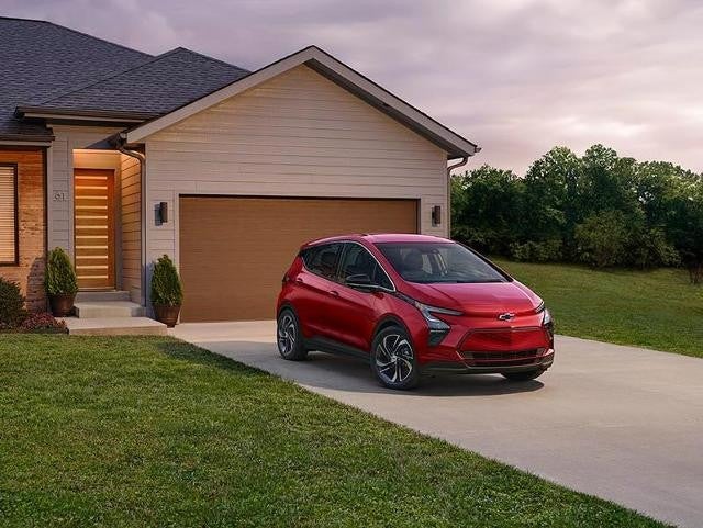 Red Chevrolet Bolt EV parked in the driveway of a suburban home during dusk, with trees and a cloudy sky in the background