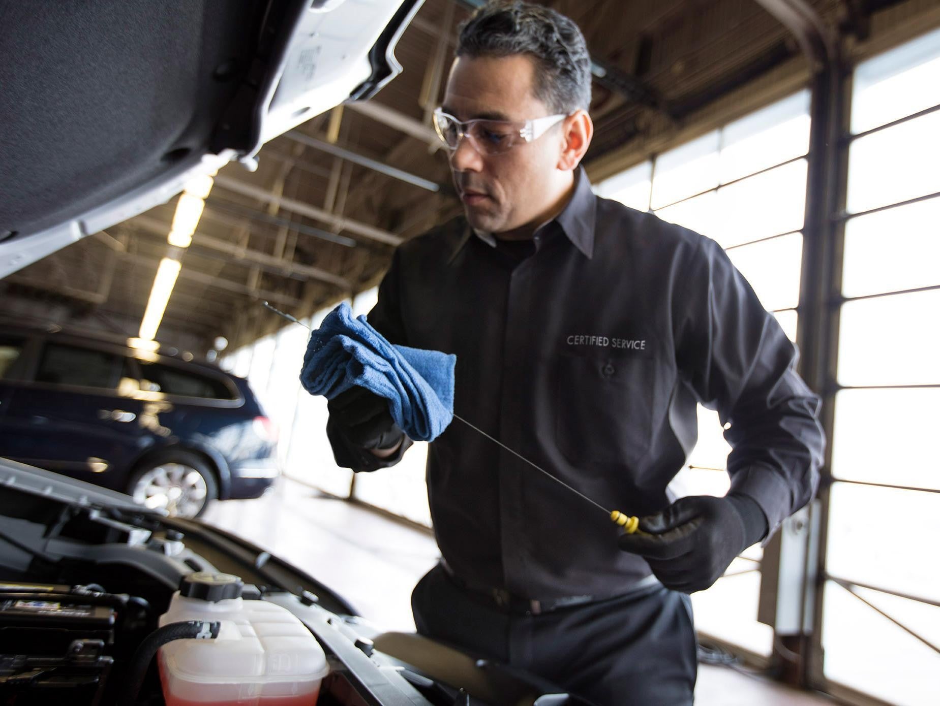 A certified Chevrolet service technician wearing safety glasses and gloves checks the engine oil level under the hood of a vehicle inside a service garage, holding a blue cloth and oil dipstick.