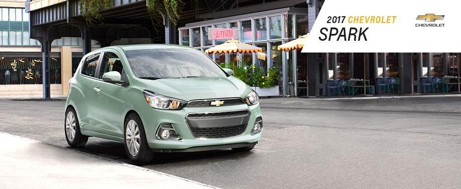 2017 Chevrolet Spark in mint green parked near a sidewalk café with outdoor seating, under city buildings with umbrellas and signage in the background.
