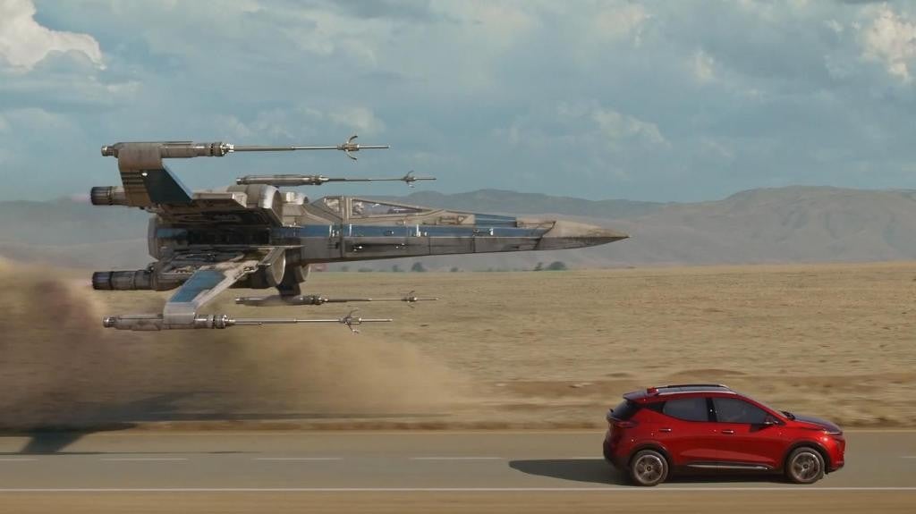 Red Chevrolet Bolt driving on a desert road beside a Star Wars X-Wing fighter in a creative promotional scene.