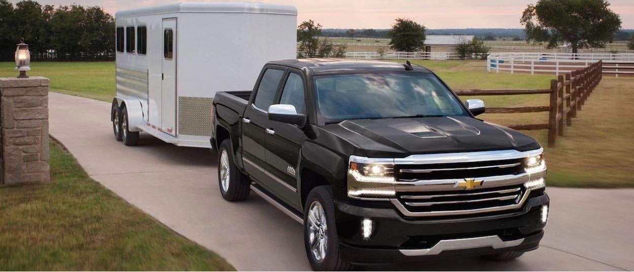 Black Chevrolet Silverado towing a white enclosed trailer on a driveway through a rural property with fences and trees in the background.